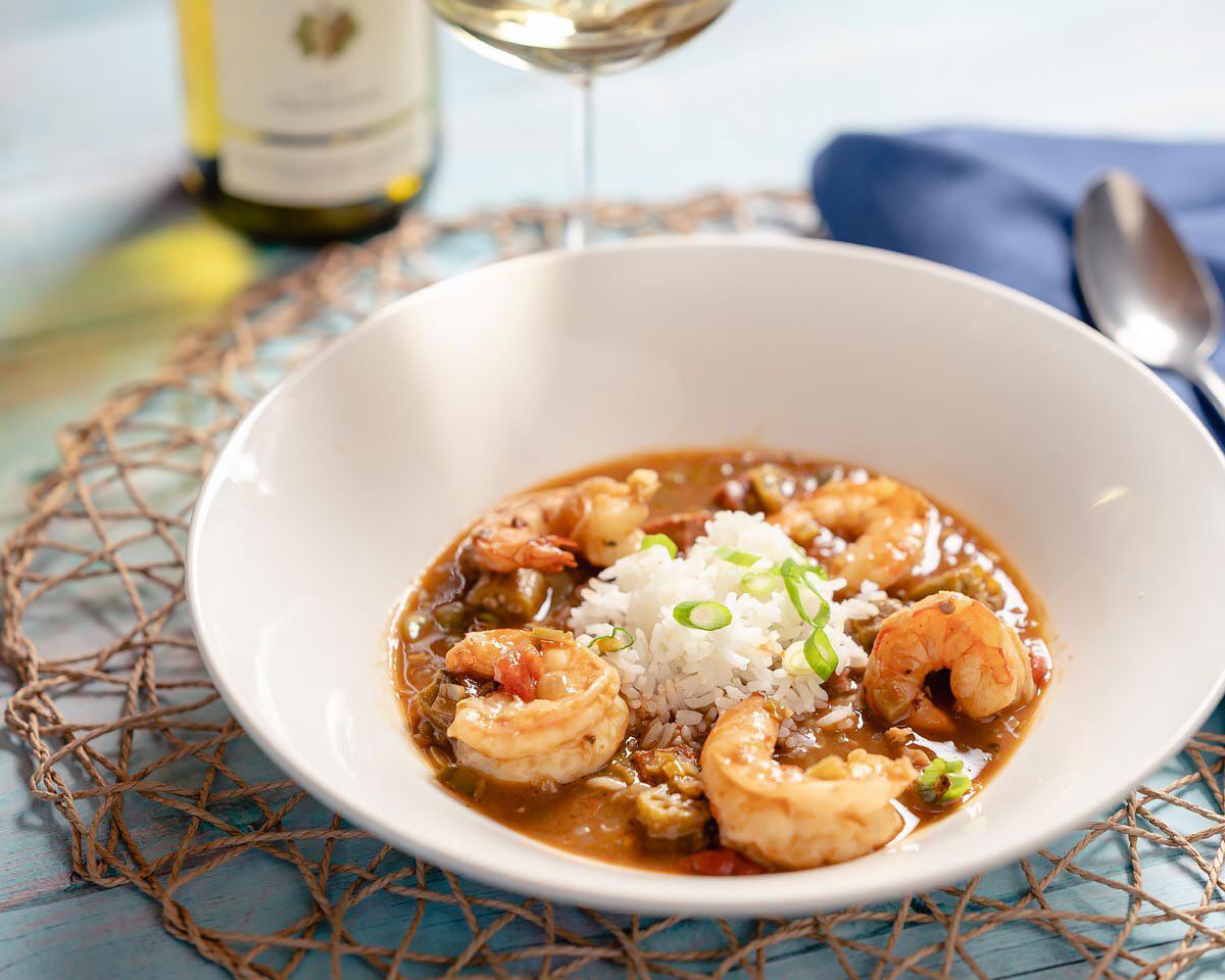 Shrimp and sausage gumbo in a white bowl, garnished with green onions, served with a glass of white wine and utensils on a bl.