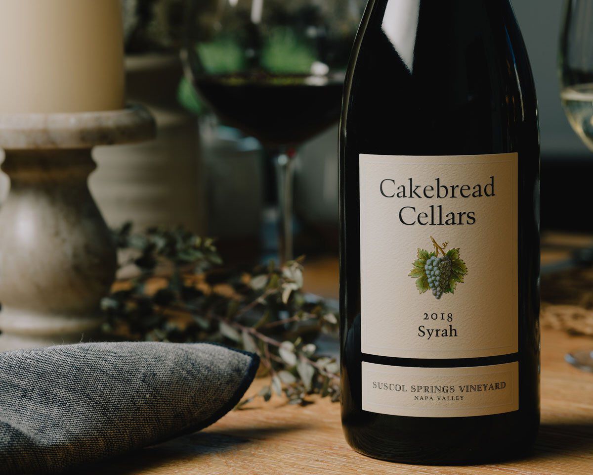 Cakebread Cellars 2018 Syrah wine bottle on a wooden table with a glass of red wine in the background.