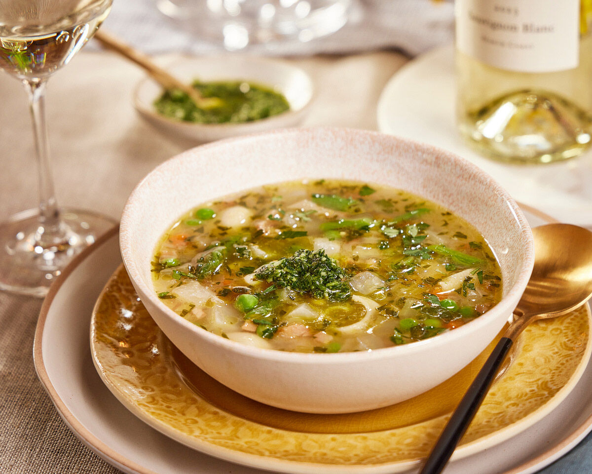 Fresh spring vegetable soup in a white bowl with green herbs and peas, served on a decorative gold-rimmed plate with a gold s.