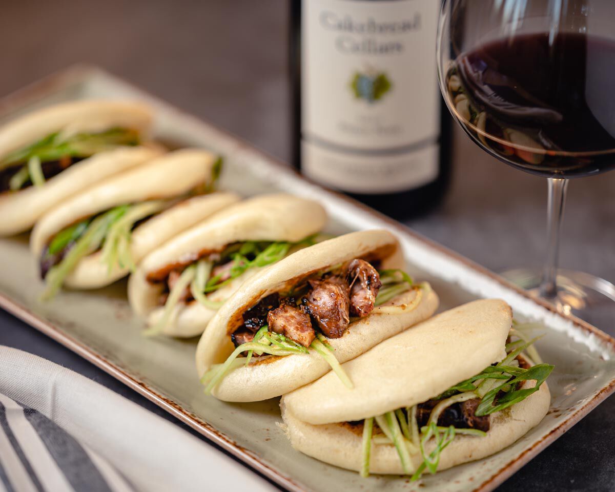 Steamed bao buns filled with Chinese BBQ pork, fresh greens, and herbs on a rectangular plate with a glass of red wine in the.