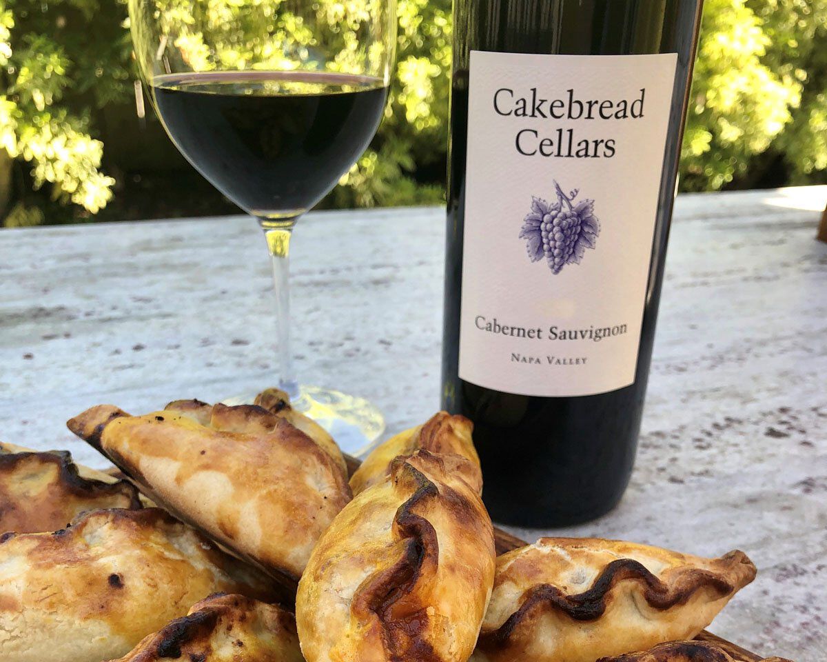 Cakebread Cellars Cabernet Sauvignon wine bottle with a glass of red wine and empanadas on a rustic table.