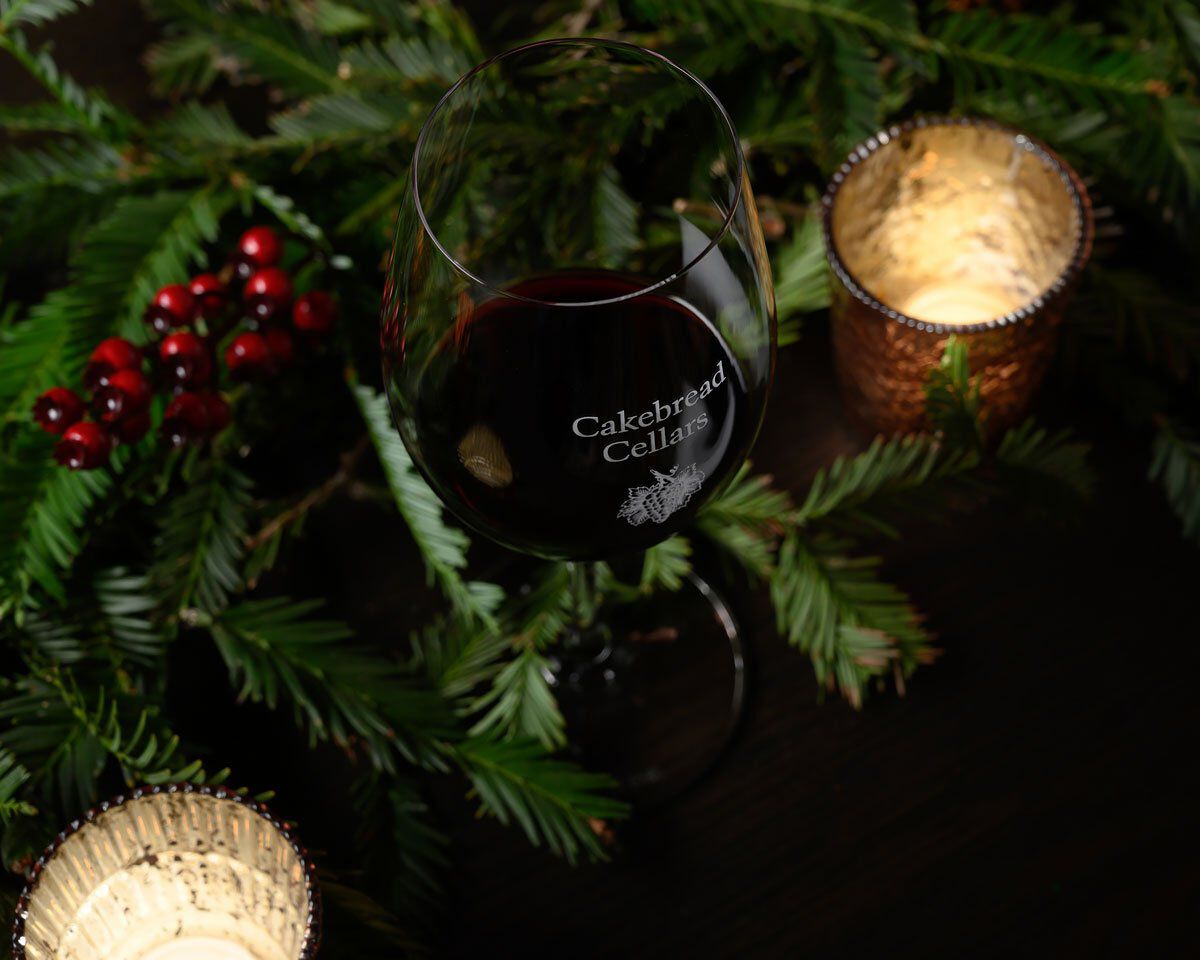 Glass of red wine with Cakebread Cellars logo, surrounded by festive greenery and candles, for a cozy holiday setting.
