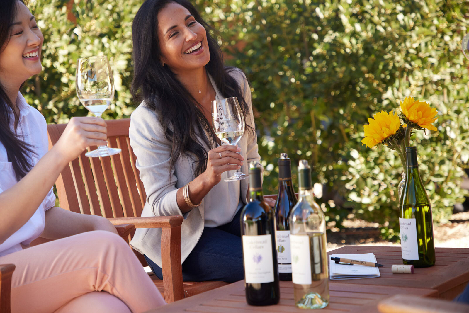 Enjoying wine outdoors with friends at a vineyard, surrounded by lush greenery and bright yellow flowers, during a sunny day.
