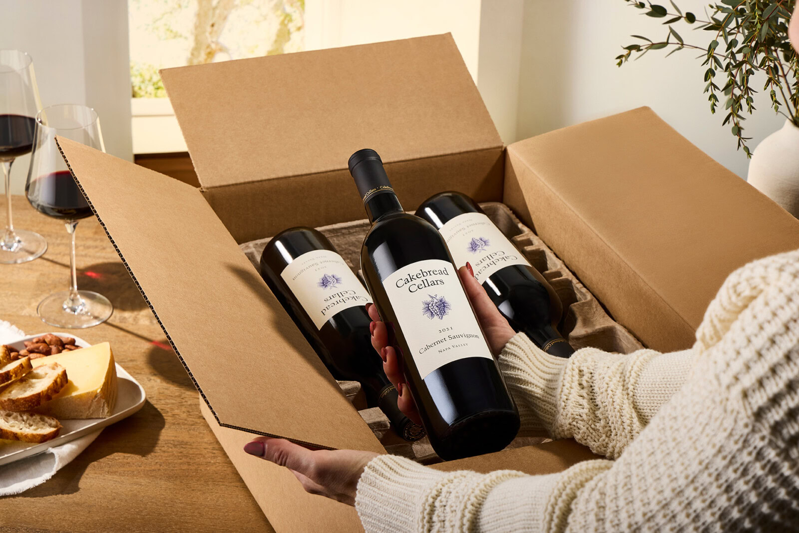A woman holding three bottles of Cakebread Cellars wine in a cardboard box.