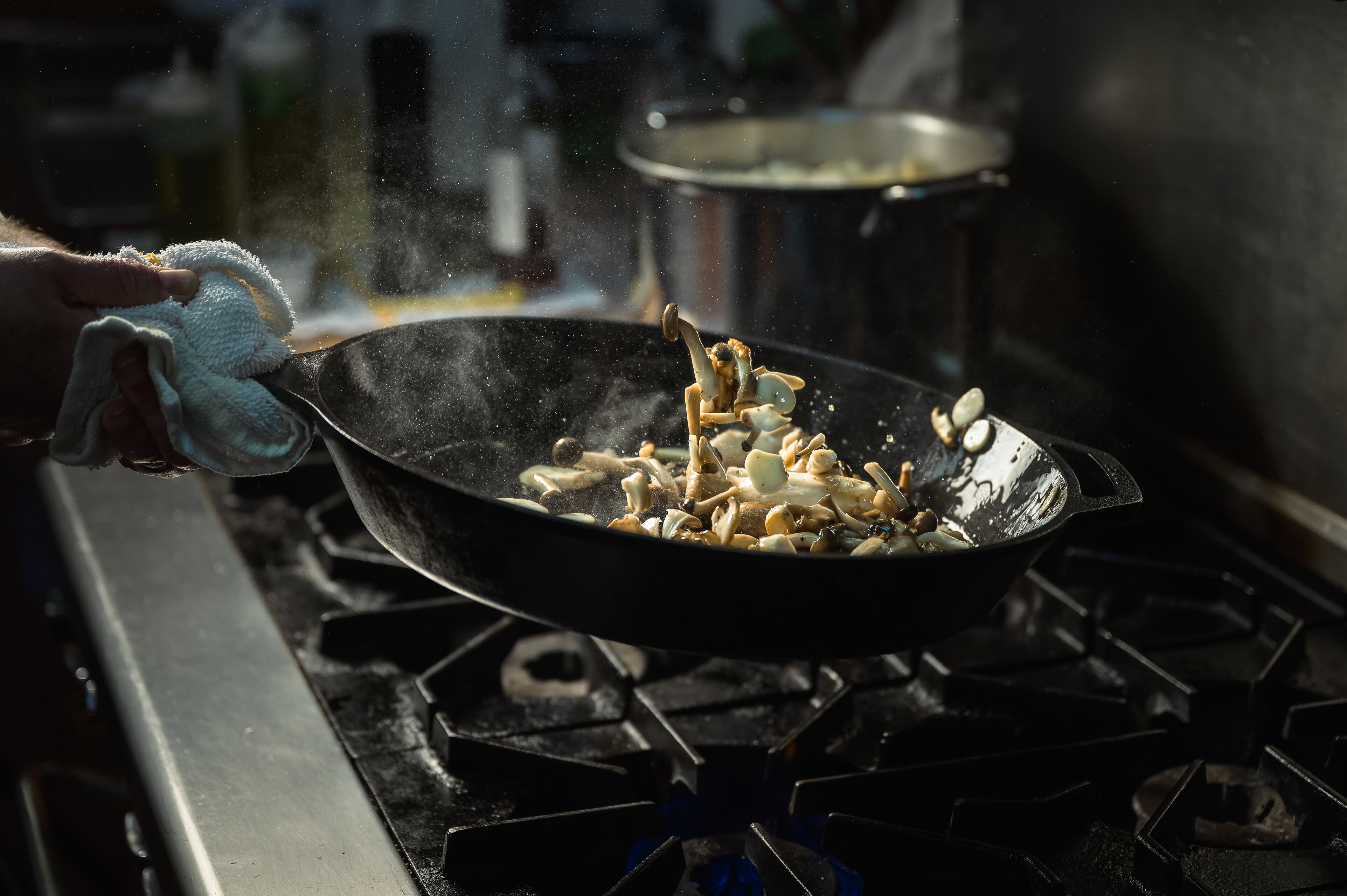 Sautéing mushrooms in a black skillet on a stove with a hand holding the skillet handle, steam rising, in a kitchen setting.