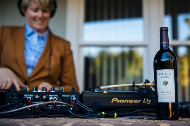 A woman DJing at a table with a bottle of Cakebread Cellars wine in the foreground.