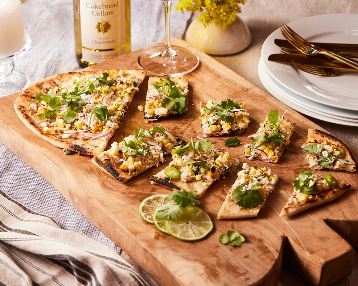 Thin-crust flatbread topped with cheese, herbs, and greens on a wooden serving board for a cooking with Cakebread feature.