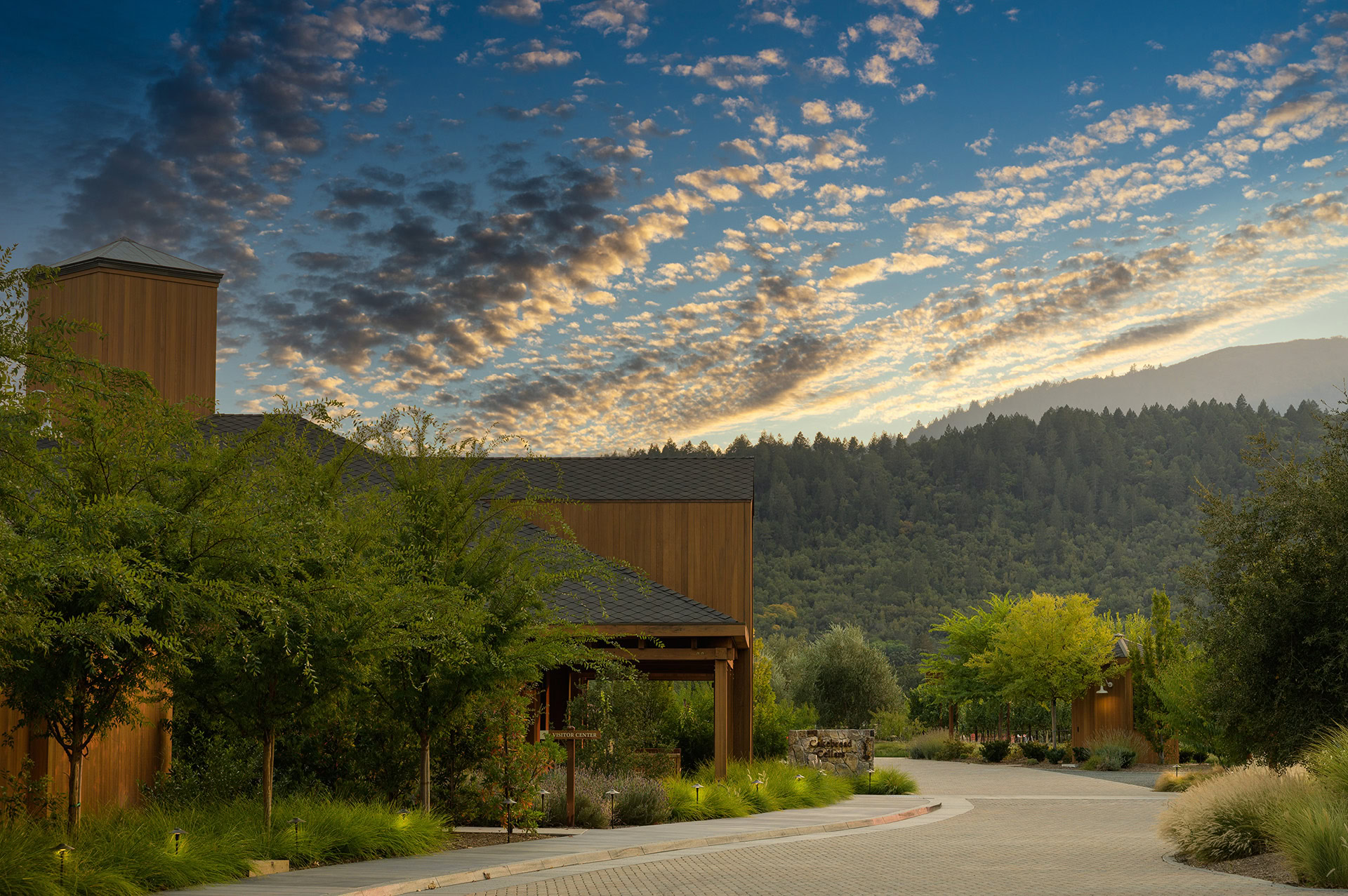 A lush landscape at sunset with modern wooden buildings, green trees, and rolling hills in the background.