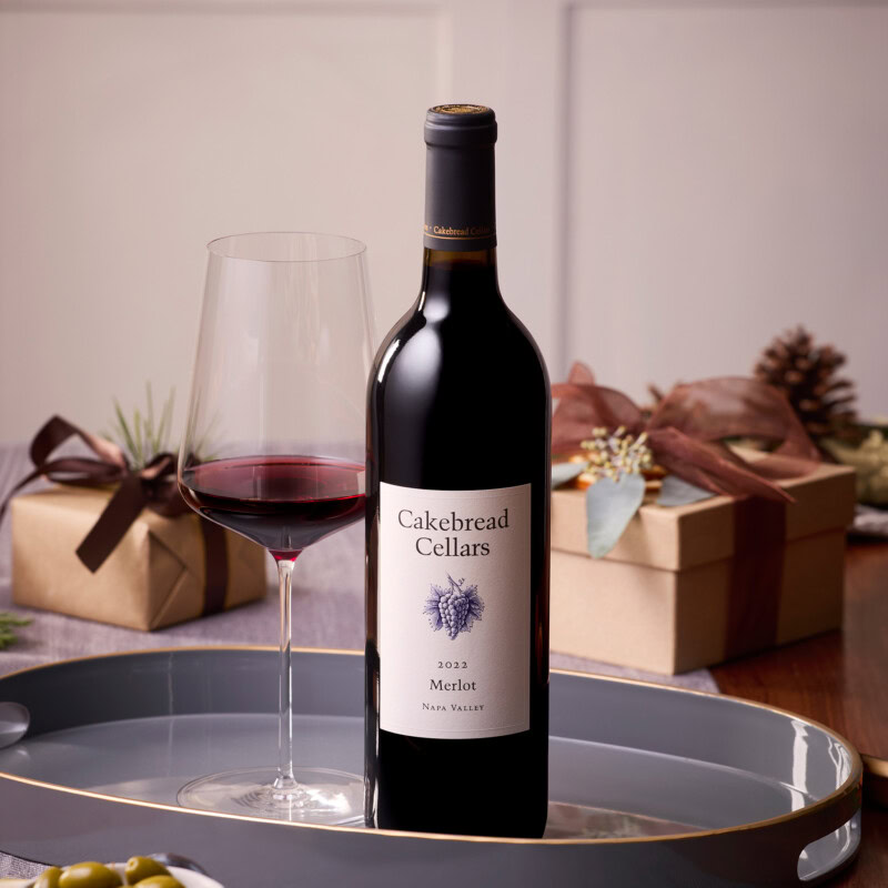 Bottle of Cakebread Cellars Merlot wine with a glass of red wine on a tray, holiday gifts in the background.