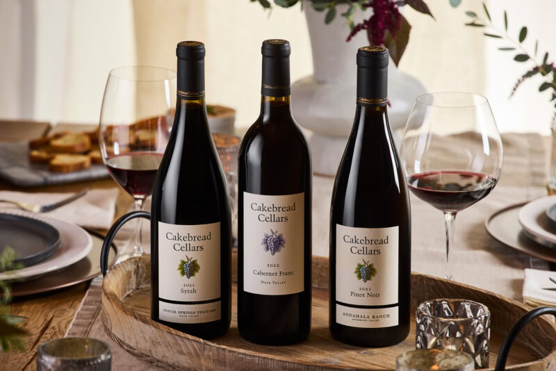 A lush display of three Cakebread Cellars wine bottles with glasses of red wine on a rustic table setting.