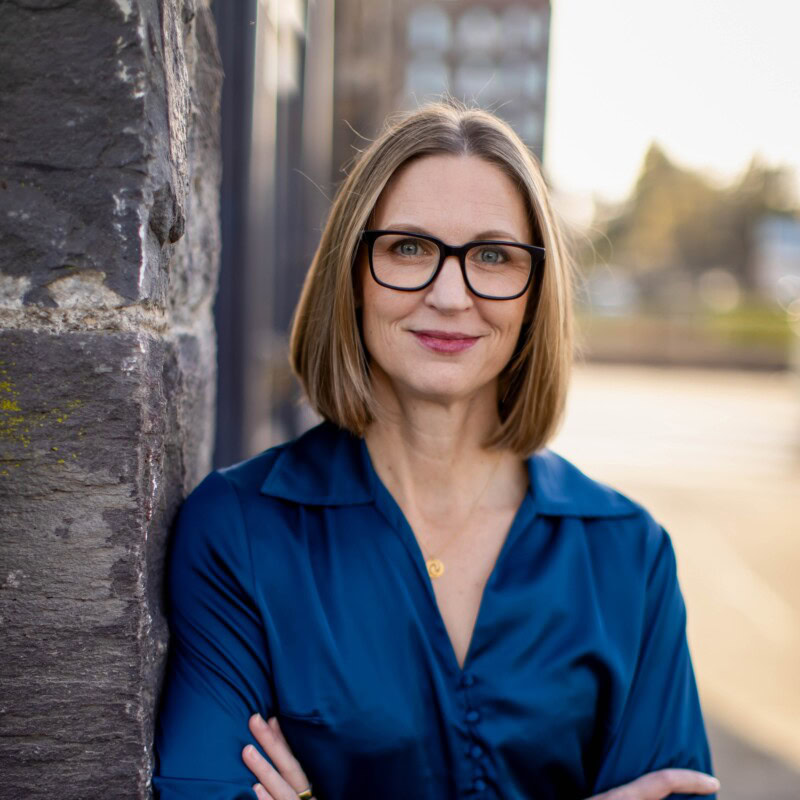 A portrait of Sara Rathbun, wearing a blue blouse, standing outdoors near a stone wall.