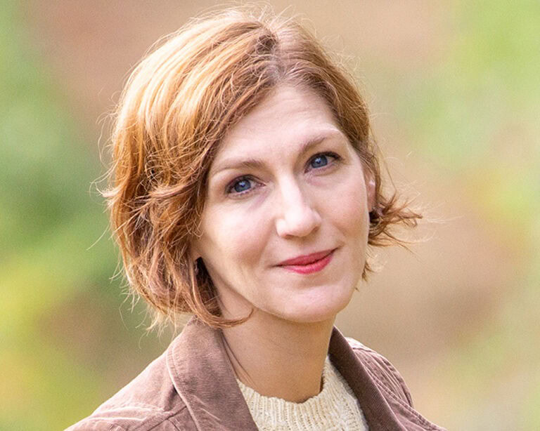A portrait of Niki Williams, posing outdoor in a light blazer and smiling at the camera.
