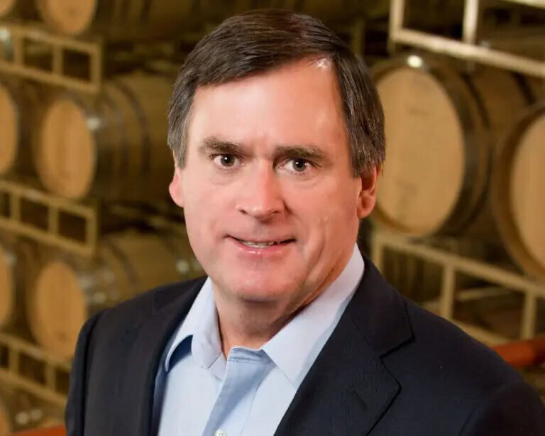 Portrait of Dennis Cakebread in a suit at Cakebread Cellars wine cellar with barrels in the background.