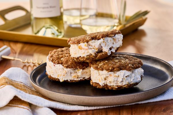 Three ice cream sandwiches, oatmeal cookies with peach ice cream on a brown plate with white wine in the background