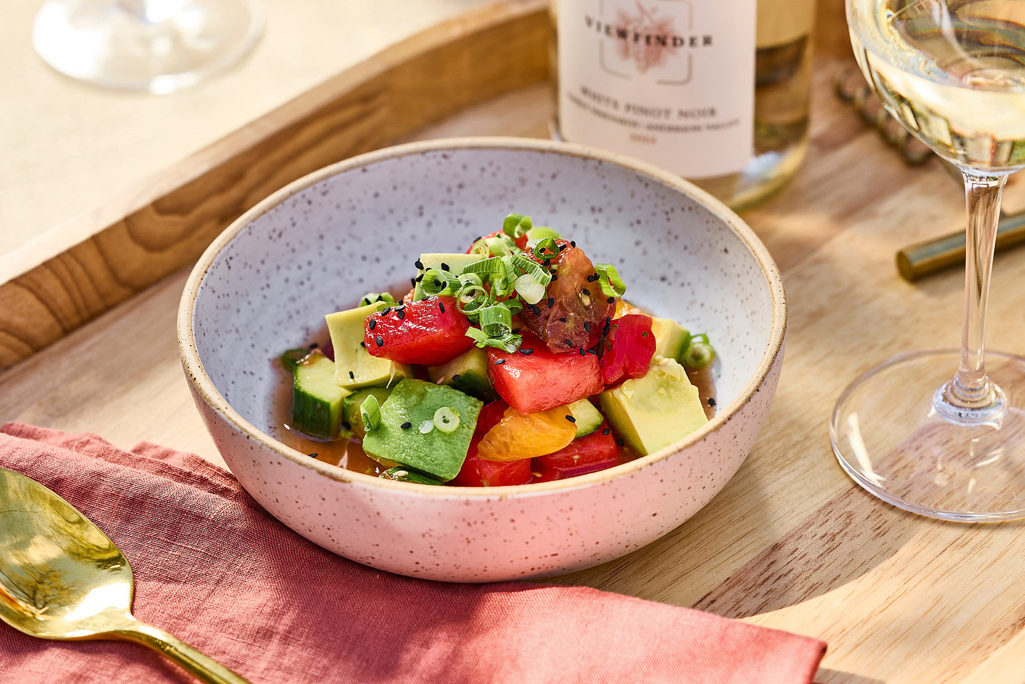 Avocado, tomatoes, cucumbers in cream speckled bowl with a soy dressing on a wood tray with a glass of wine.
