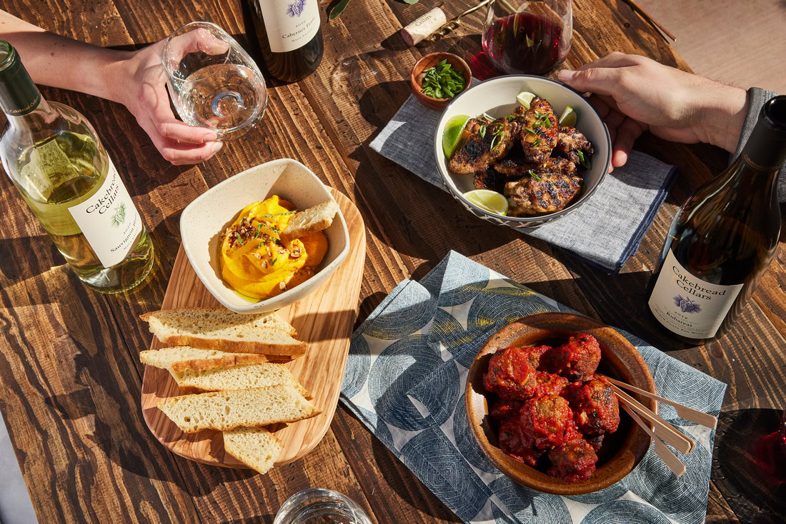 Wine and food tasting with bottles of Cakebread Cellars wine, cheese, and appetizers on a rustic wooden table.