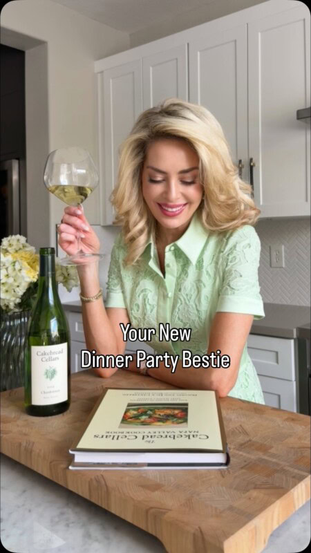 A woman smiling and holding a glass of white wine in a modern kitchen with a bottle of Cakebread Cellars wine on the counter.