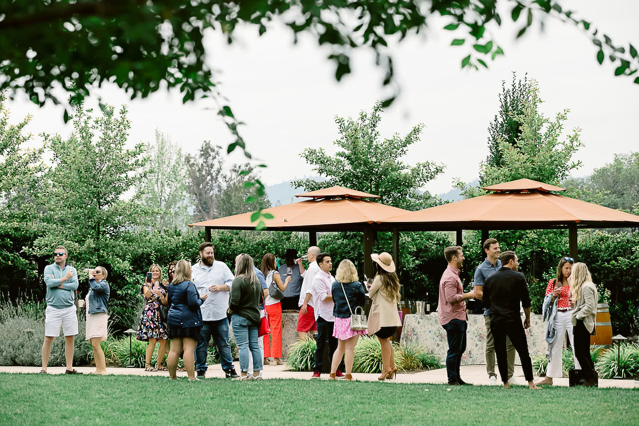 Group of people enjoying wine tasting outdoors at Cakebread Cellars with lush greenery and shaded umbrellas.
